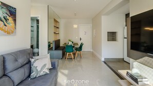A modern living room with a grey sofa, a dining area with a table and chairs, and a kitchen area with a TV and oven.