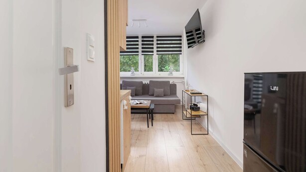 A modern living room with a grey sofa, a wooden dining table, and a black refrigerator. - Comfortable Apartment | Balcony | Close to Silesian Park (Katowice)