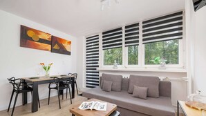 A modern living room with a grey sofa, a dining table with chairs, and a window with blinds. - Comfortable Apartment | Balcony | Close to Silesian Park (Katowice)