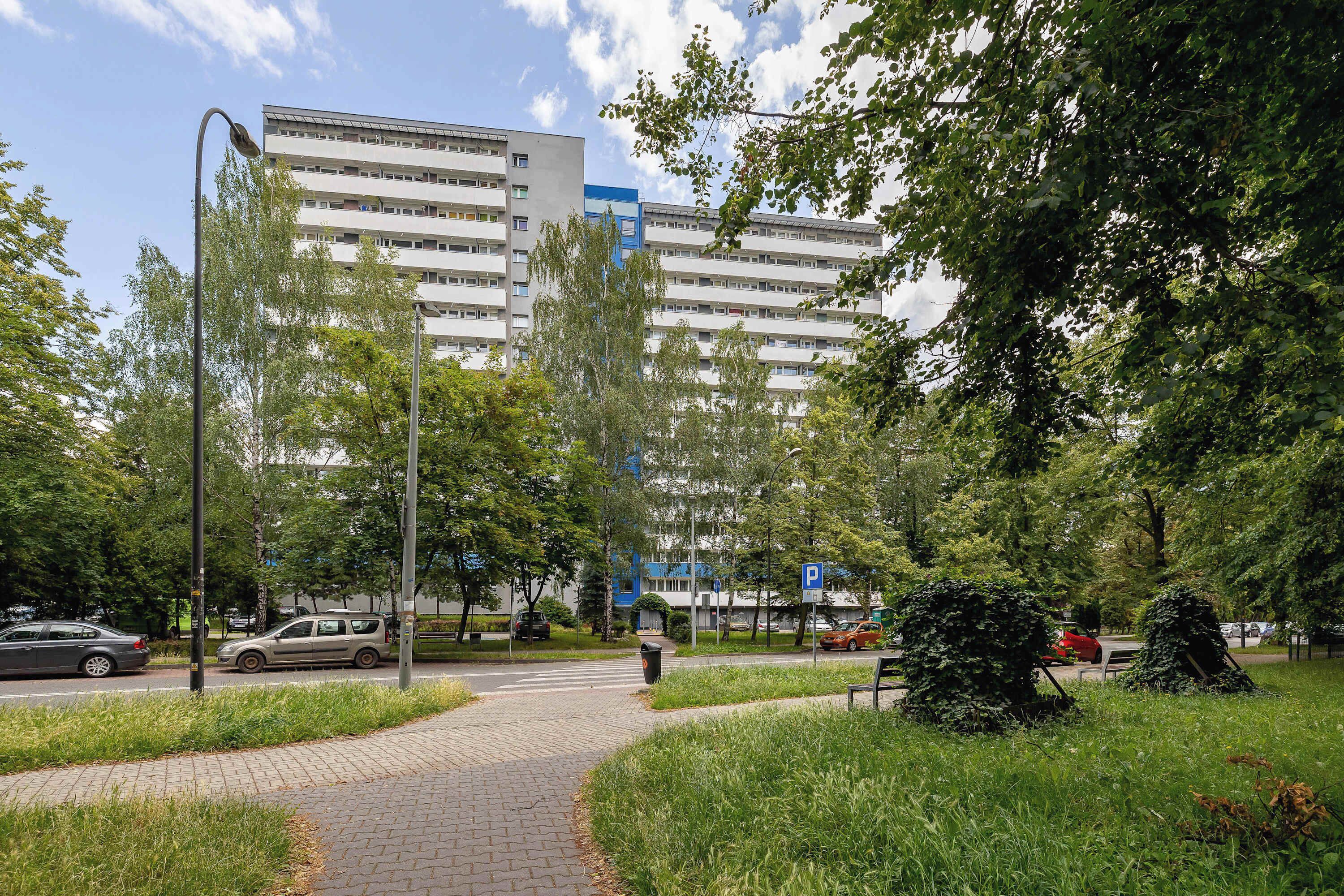 Un sendero pavimentado con césped y árboles, que conduce a un edificio de varios pisos con una sección azul.