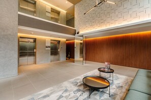 A modern lobby with a glass elevator, a wooden panel wall, and a seating area with a patterned rug.