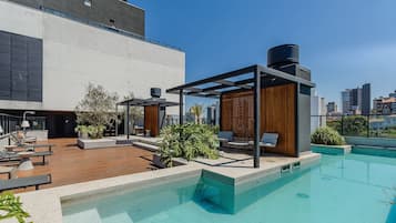 A rooftop pool area with lounge chairs, a pergola, and a view of the city skyline.