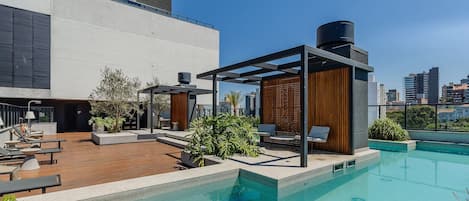 A rooftop pool area with lounge chairs, a pergola, and a view of the city skyline.