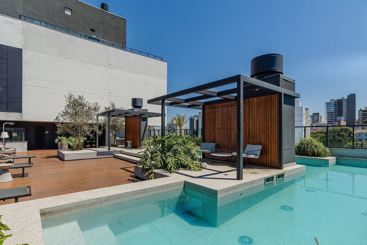 A rooftop pool area with lounge chairs, a pergola, and a view of the city skyline.