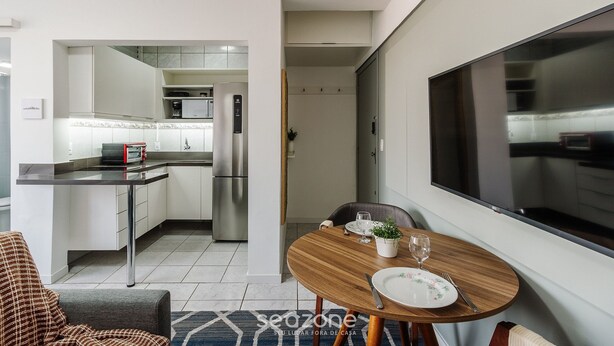A modern kitchen with white cabinets, a stainless steel refrigerator, and a built-in oven. There is a dining area with a round wooden table, a white plate, and a glass of water. - Practical Studio Near Farroupilha Park Hpn707 (Porto Alegre)