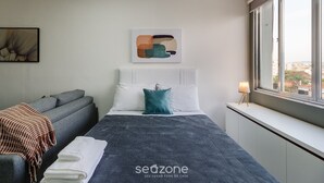 A modern hotel room with a bed, a sofa, a window with a city view, and a wall art piece. - Practical Studio Near Farroupilha Park Hpn707 (Porto Alegre)