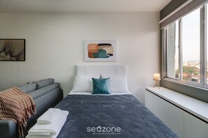 A modern hotel room with a bed, a sofa, a window with a city view, and a wall art piece.