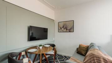 A modern living room with a flat-screen TV mounted on the wall, a round dining table with chairs, a sofa, and a patterned rug.