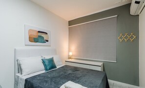 Un dormitorio moderno con cama, almohadas, mesita de noche con lámpara y aire acondicionado de pared.