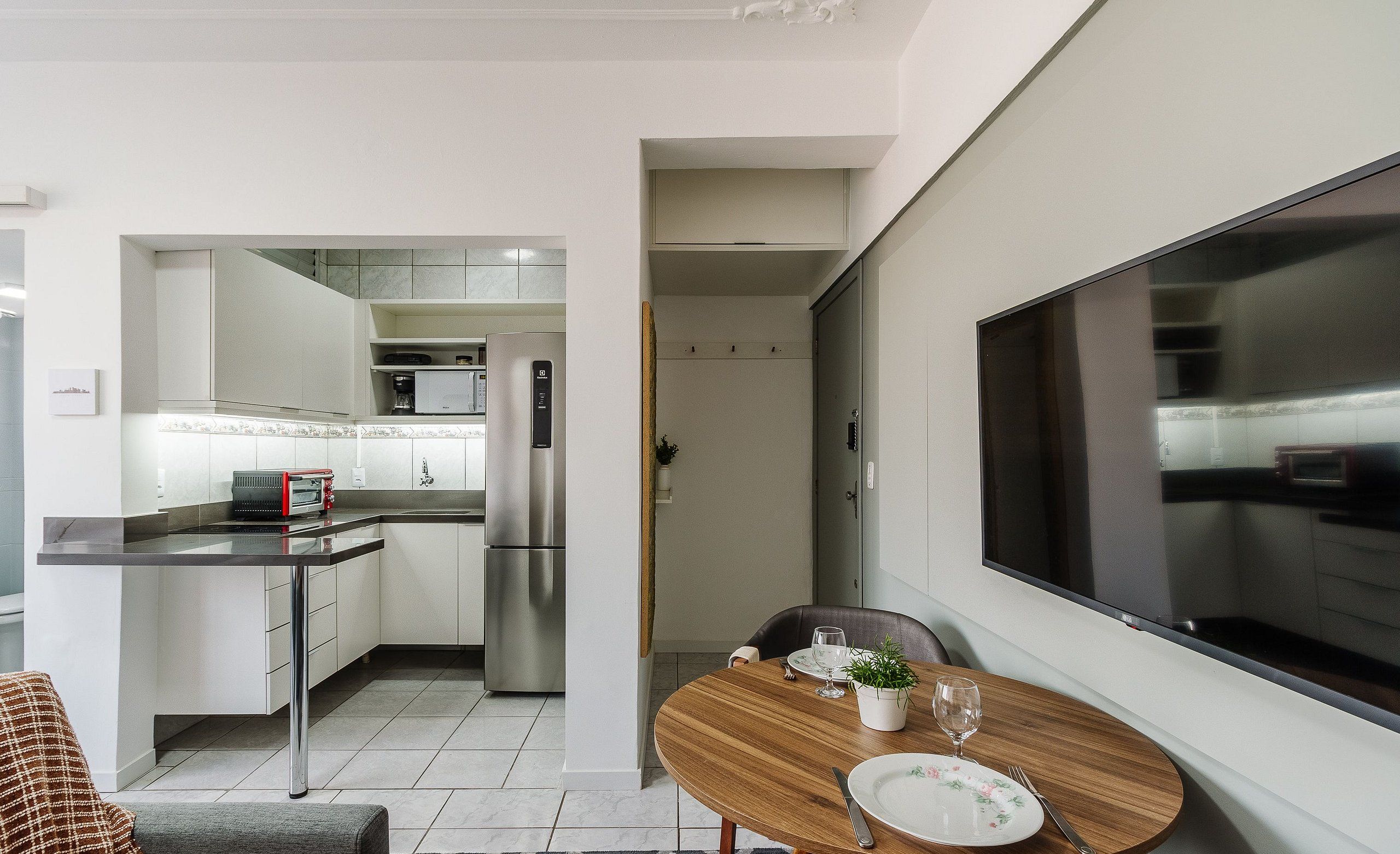 A modern kitchen with white cabinets, a stainless steel refrigerator, and a built-in oven. There is a dining area with a round wooden table, a white plate, and a glass of water.