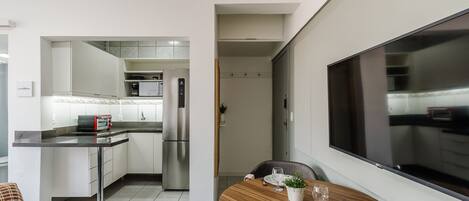 A modern kitchen with white cabinets, a stainless steel refrigerator, and a built-in oven. There is a dining area with a round wooden table, a white plate, and a glass of water.