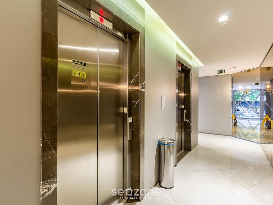 Un ascensor moderno con un cartel que indica atención a la accesibilidad, un cubo de basura y un pasillo que conduce a un espacio con paredes de vidrio.