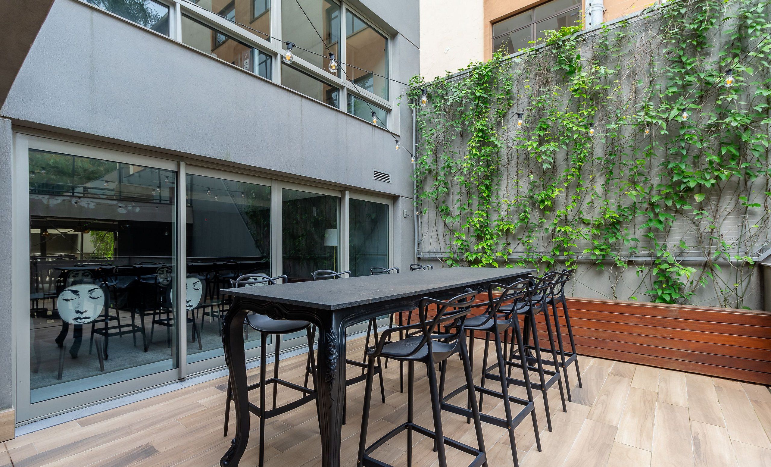 A modern outdoor dining area with a long table and chairs, a glass partition, and a wall covered in climbing plants.