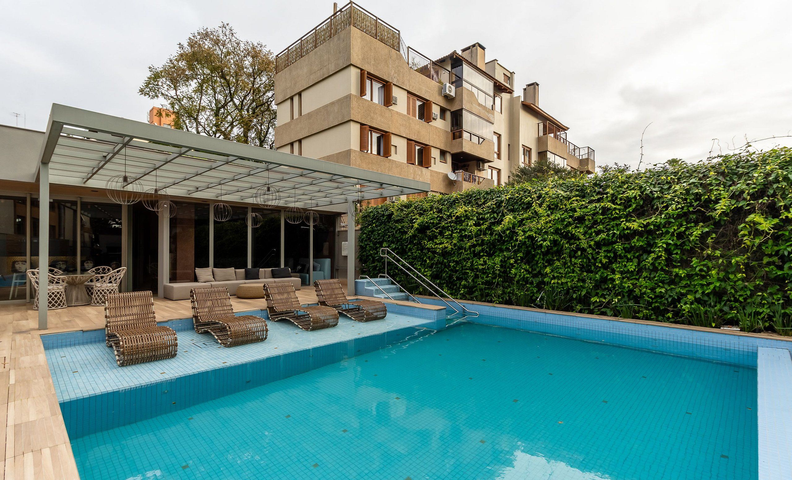 Piscina con sillas de descanso y una zona de estar cubierta, rodeada de un seto y un edificio de varios pisos al fondo.