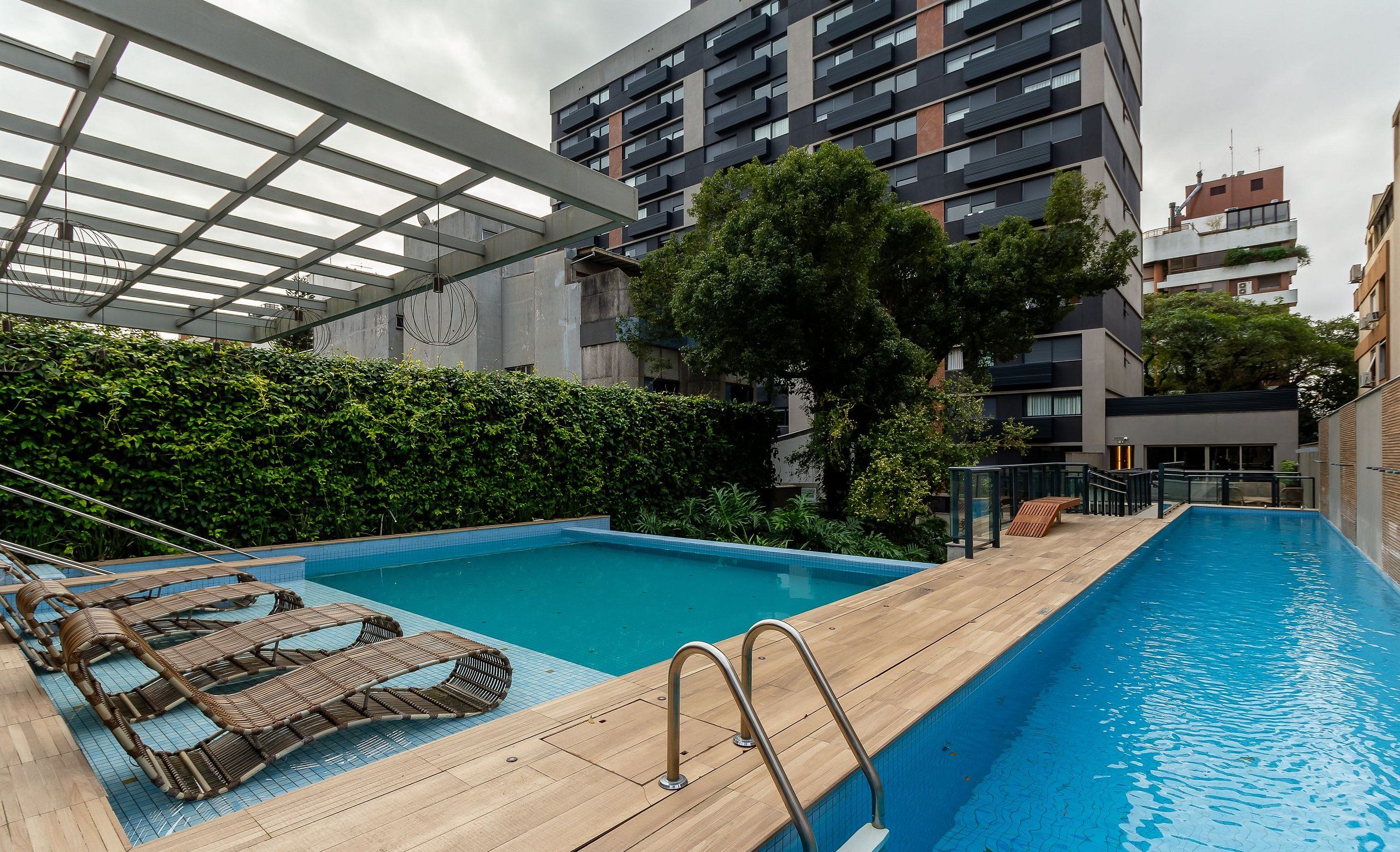Una piscina en la azotea con tumbonas de madera y escaleras de metal, rodeada de vegetación y edificios modernos.