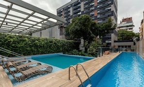 Une piscine sur le toit, dotée de chaises longues en bois et d’échelles en métal, entourée de verdure et de bâtiments modernes.