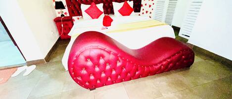 A bed with a red headboard and white bedding, a red upholstered bench, and a tiled floor.