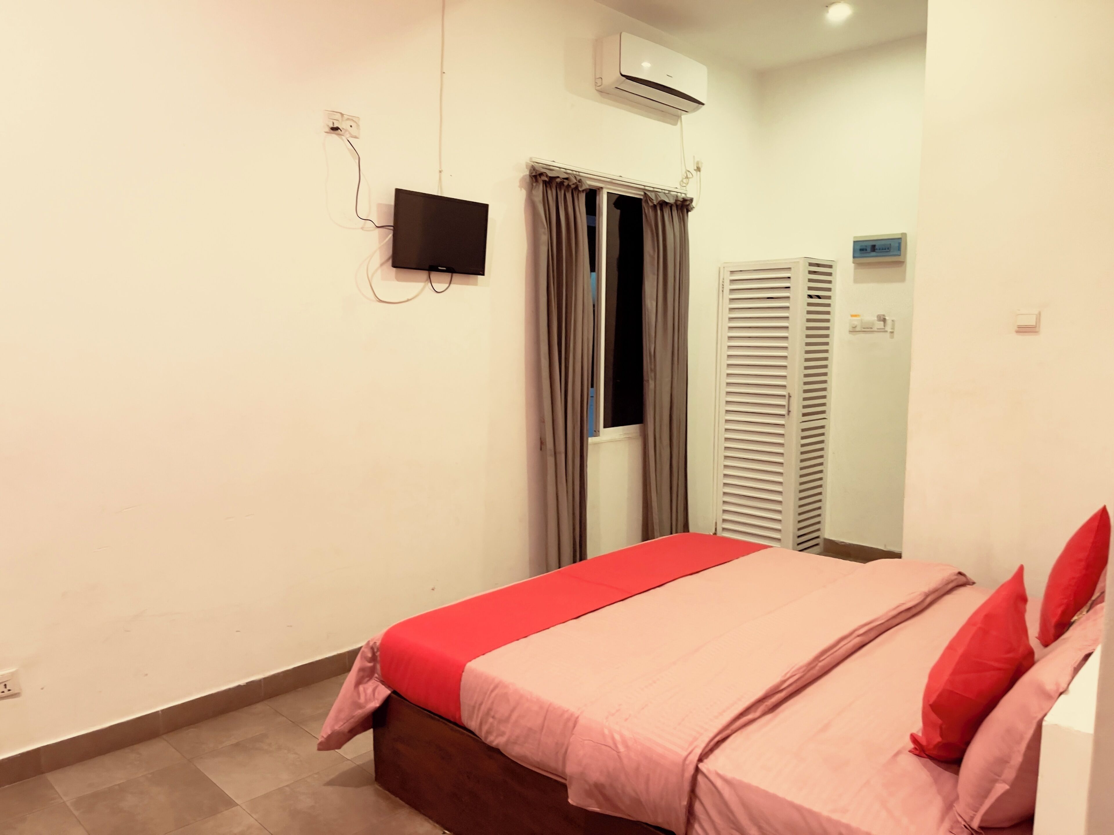 A bedroom with a bed, a TV mounted on the wall, an air conditioner, and a window with curtains.