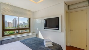 A hotel room with a bed, a TV mounted on the wall, a window with blinds, and a radiator under the window. - Comfort and a Beautiful View! Pgr1101 (Porto Alegre)