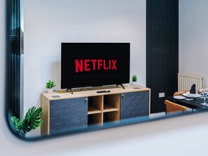 Una sala de estar moderna con un televisor de pantalla plana montado en un mueble de madera, una zona de comedor con una mesa preparada para dos personas y una planta en maceta sobre el mueble.