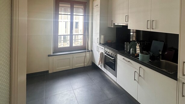Cocina compacta con armarios blancos, horno integrado y ventana con persianas.