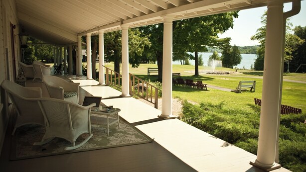 A covered porch with white columns, wicker chairs, and a view of a lake and park. - Fountain Point - Hotel Room 1 (Lake Leelanau)