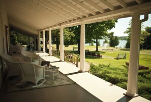 A covered porch with white columns, wicker chairs, and a view of a lake and park.