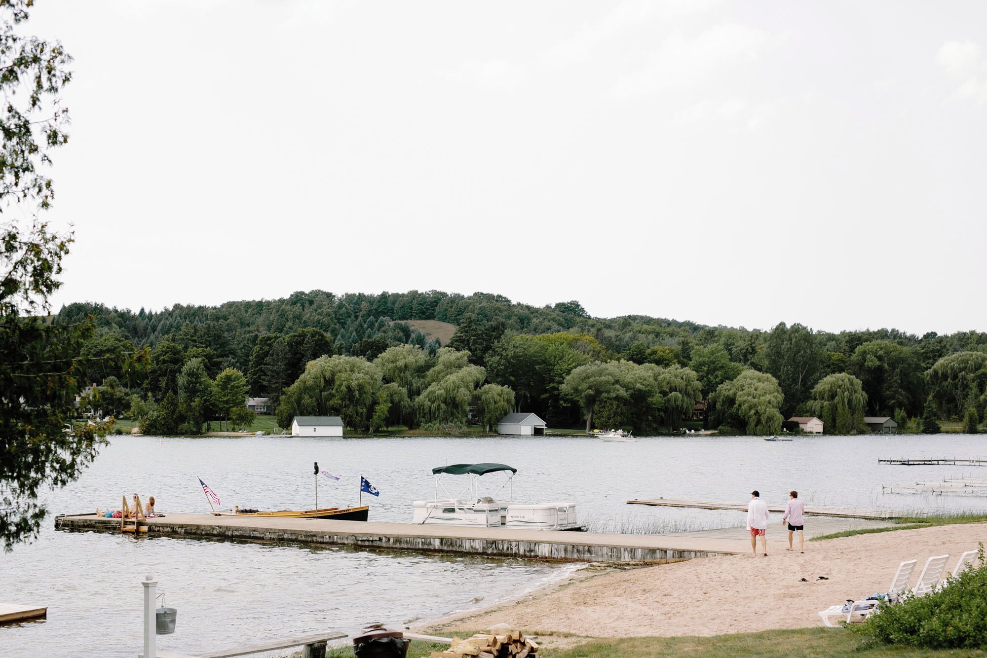 A lakeside dock with boats, a sandy beach, and a forested hill in the background.