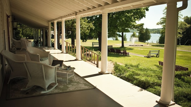 A covered porch with white columns, wicker chairs, and a view of a lake and park. - Fountain Point - Hotel Room 6 (Lake Leelanau)