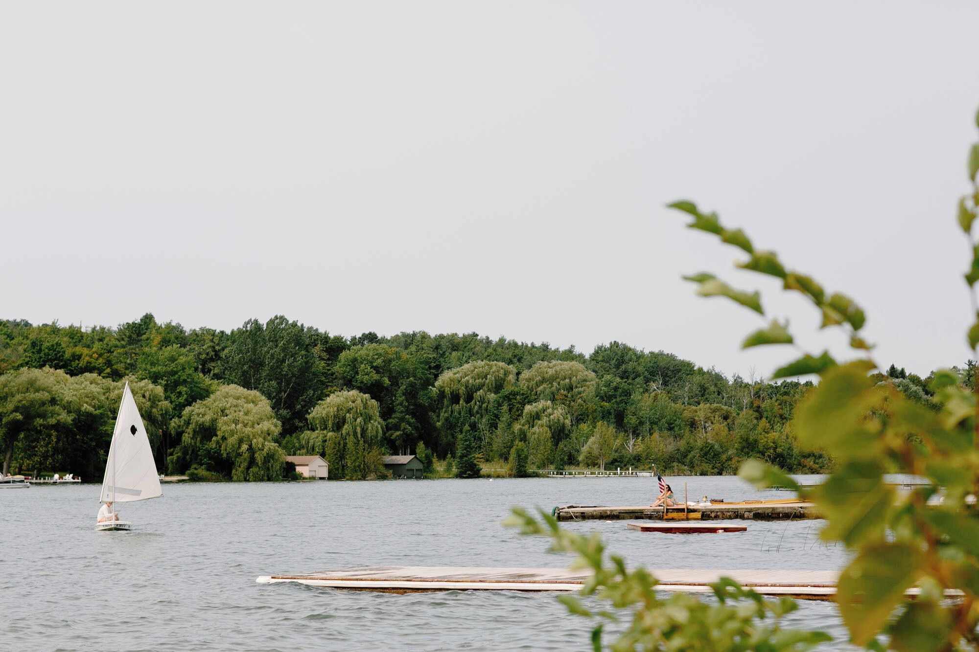 A lake with a sailboat and docked boats, surrounded by dense trees and a few buildings in the background.