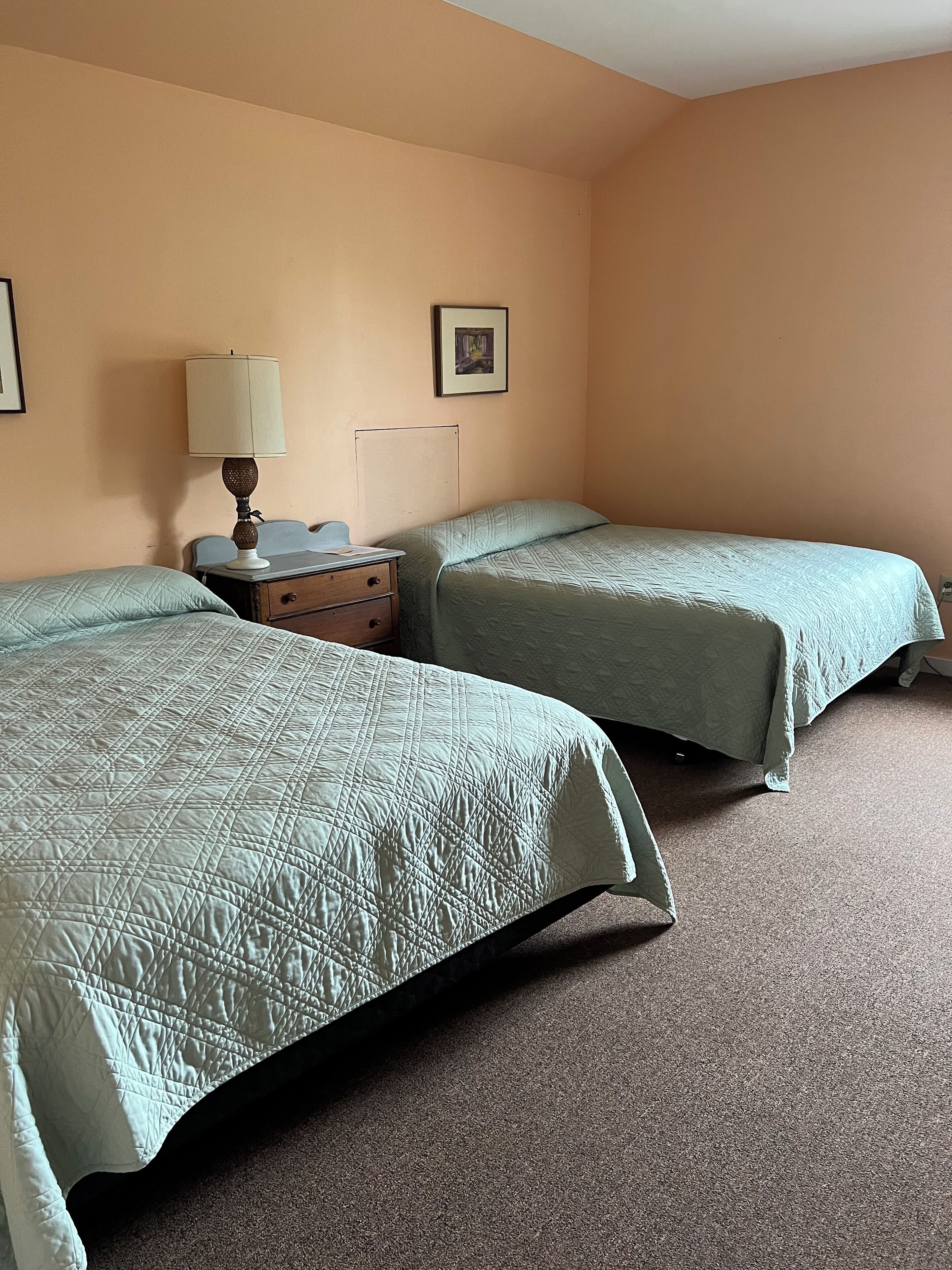 A hotel room with two beds, a nightstand, a lamp, and a framed picture on the wall.