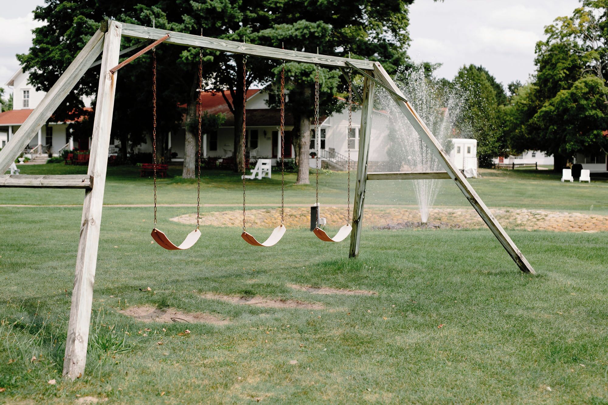 Un ensemble de balançoires en bois installé dans un espace verdoyant d’un parc, avec une fontaine en arrière-plan.