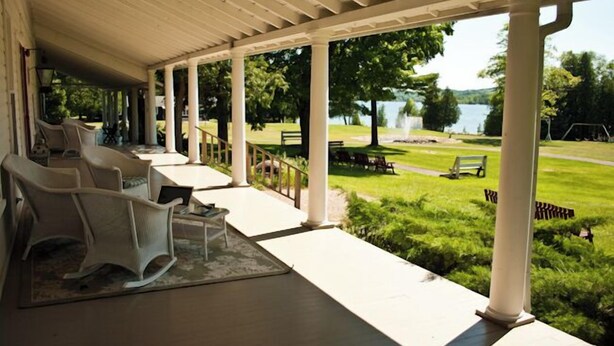 Eine überdachte Veranda mit weißen Säulen, Gartenmöbeln und Blick auf einen See und einen Park.