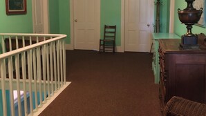A hallway with a white railing, a wicker chair with a red cushion, and a lamp on a wooden table. - Fountain Point - Hotel Room 17 (Lake Leelanau)