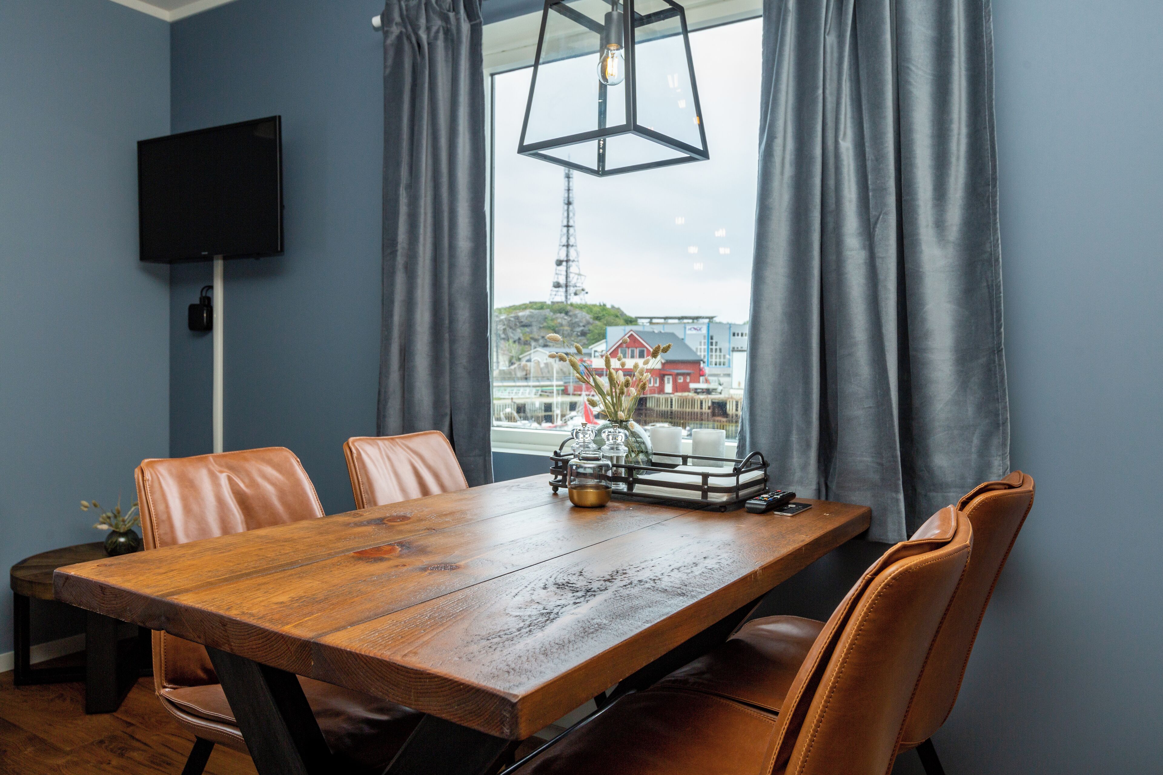 A dining area with a wooden table, leather chairs, a TV mounted on the wall, and a view of a building through a window with curtains.