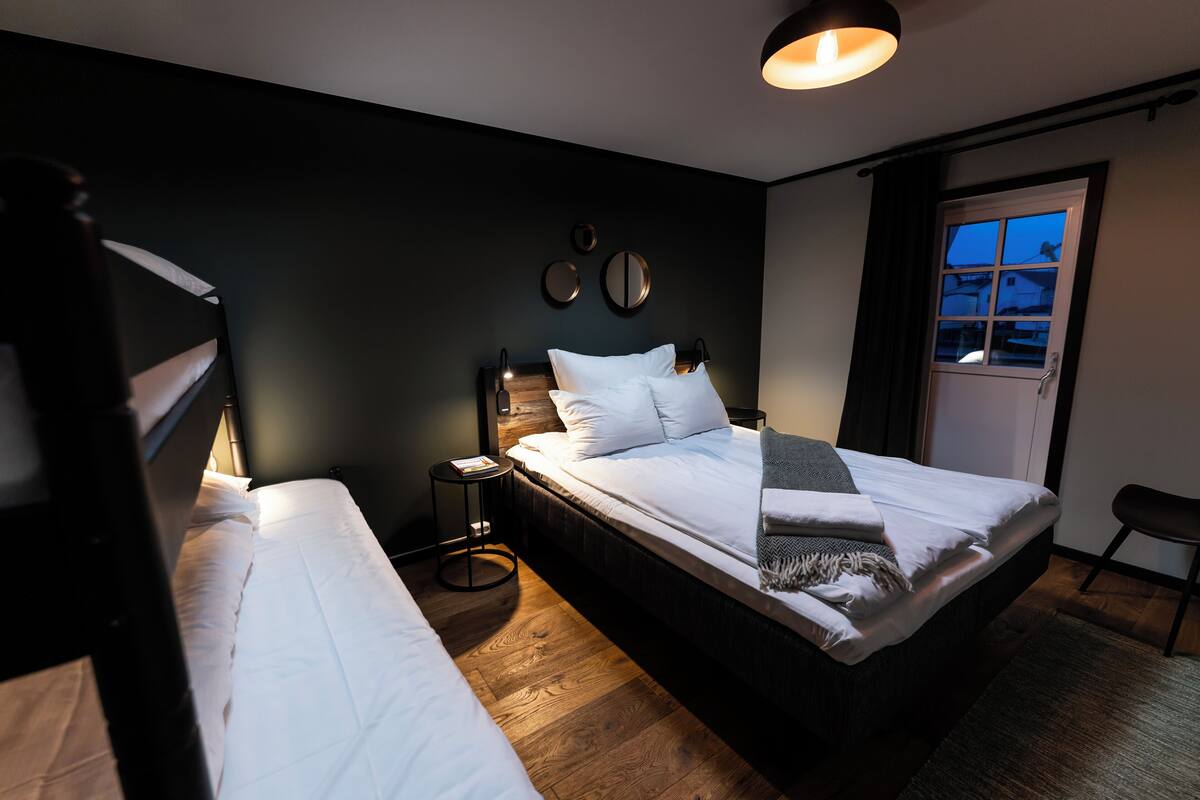 A hotel room with two beds, a small bedside table, a round mirror, and a window with curtains.
