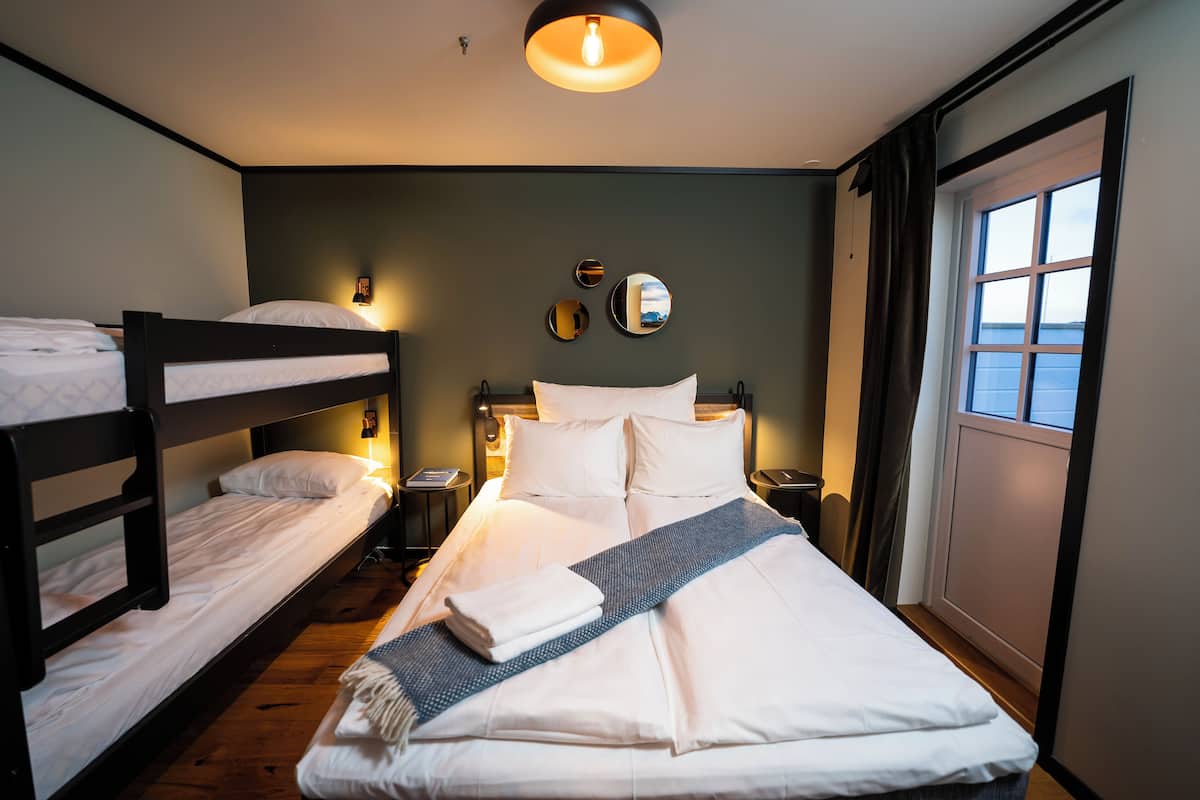 A hotel room with a bunk bed, a single bed, a bedside table, a lamp, a mirror, and a window with curtains.