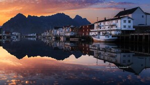 Un pueblo frente al mar con edificios reflejándose en aguas tranquilas, una cadena montañosa al fondo y un cielo despejado.