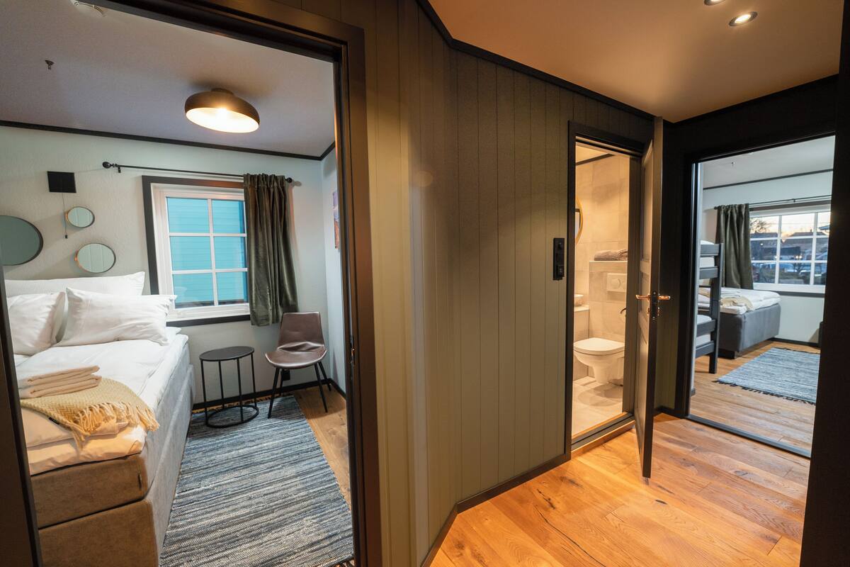 A modern hotel room with a bed, a small round table, a chair, a bathroom with a toilet and shower, and a view through a glass door.