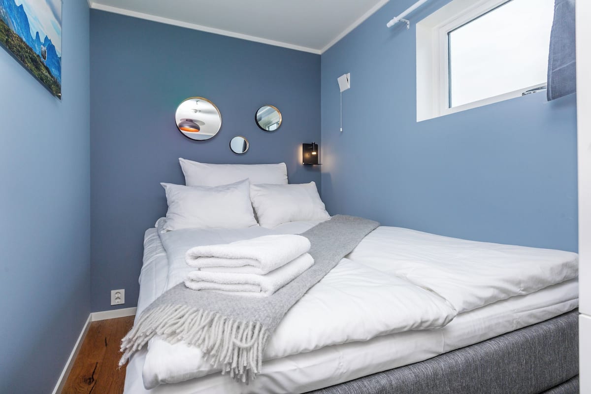 A neatly made bed with white linens and a grey throw blanket, a wall-mounted mirror, and a window with a curtain.