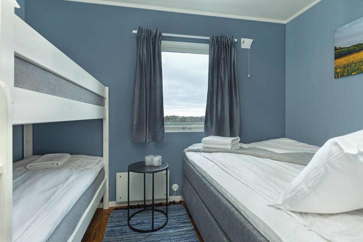 A room with two bunk beds, a small round table with a cup and tissues, a window with curtains, and a painting on the wall.