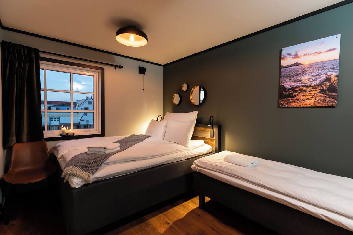 A bedroom with two beds, a window with curtains, a wall-mounted mirror, and a painting of a sunset.