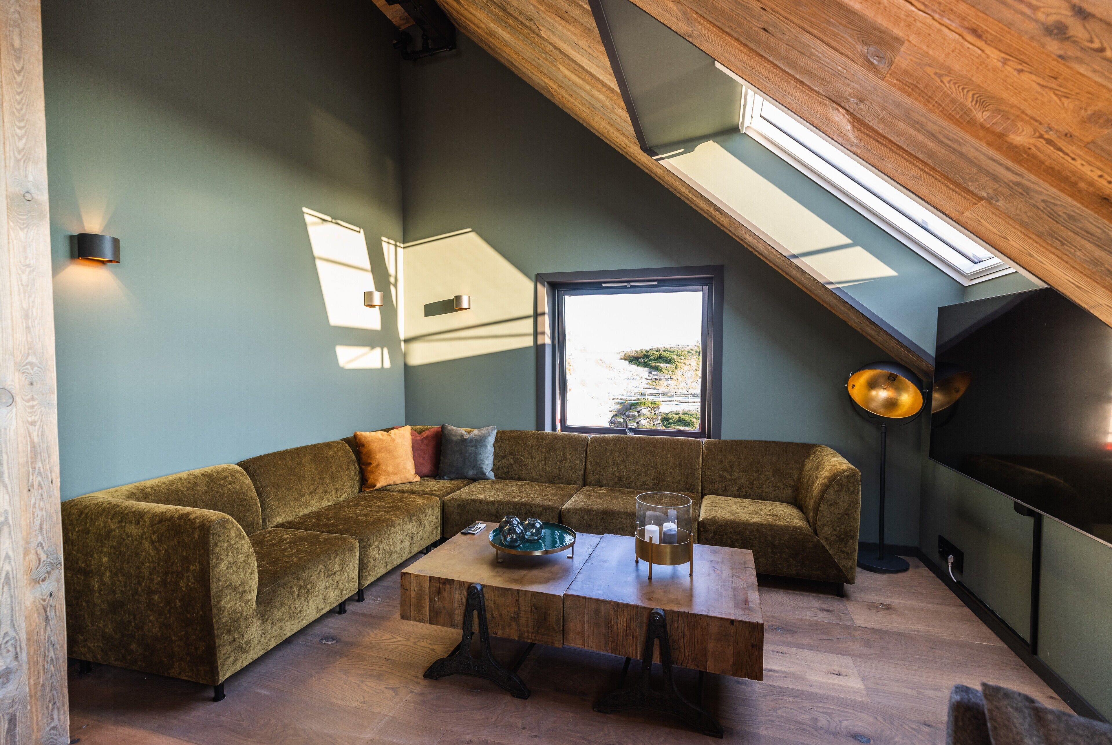 Ein Loft-Wohnraum mit Holzbalkendecke, großem Fenster, Ecksofa, Couchtisch und Stehlampe.