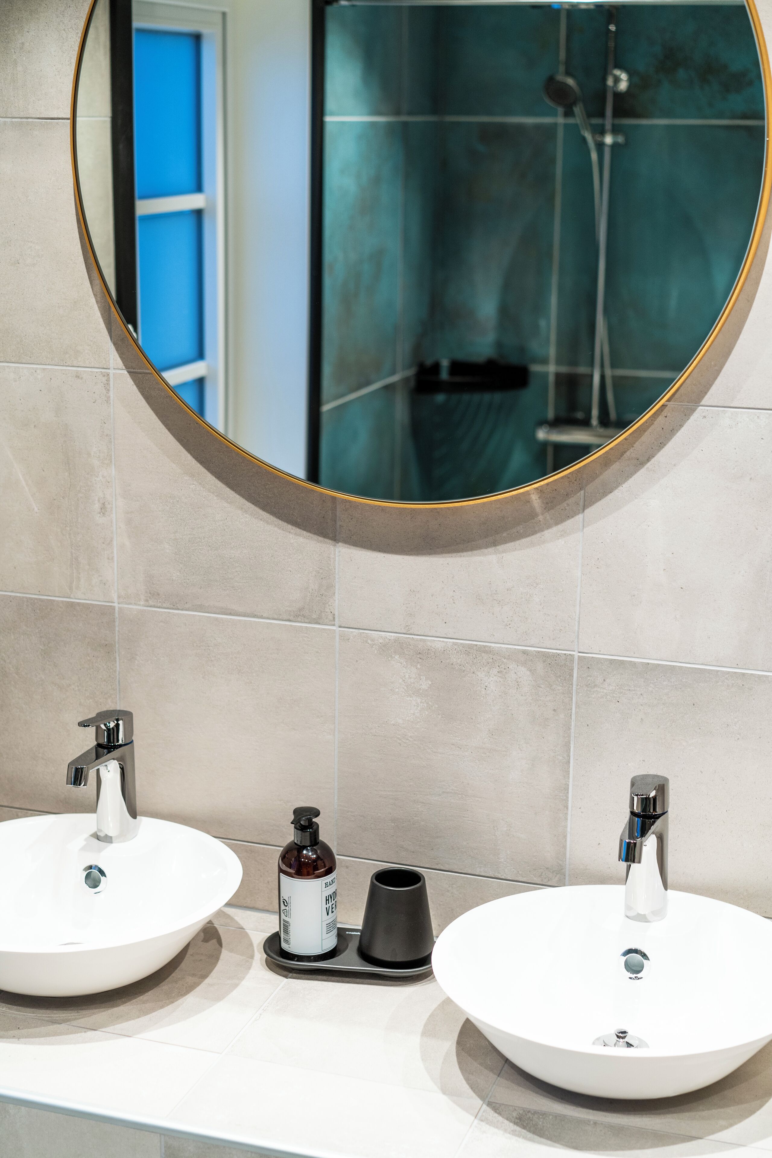 A bathroom with two white sinks, a round mirror, and a shower visible in the reflection.
