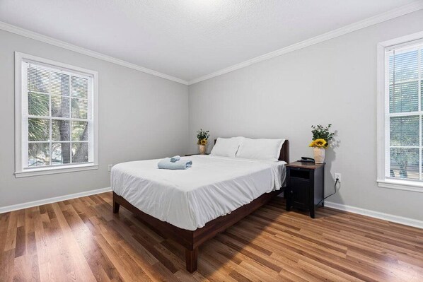 A bedroom with a wooden floor, a bed with white bedding, two bedside tables with lamps, and windows with blinds.