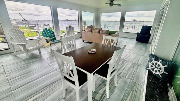 A spacious room with a large table and white chairs, a sofa, and a view of the water through large windows. - Orange Bay Retreat - Amazing View (Fort Myers Beach)