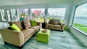 A modern living room with a sofa, a chair, and a small ottoman. There are large windows offering a view of a marina and buildings. - Orange Bay Retreat - Amazing View (Fort Myers Beach)