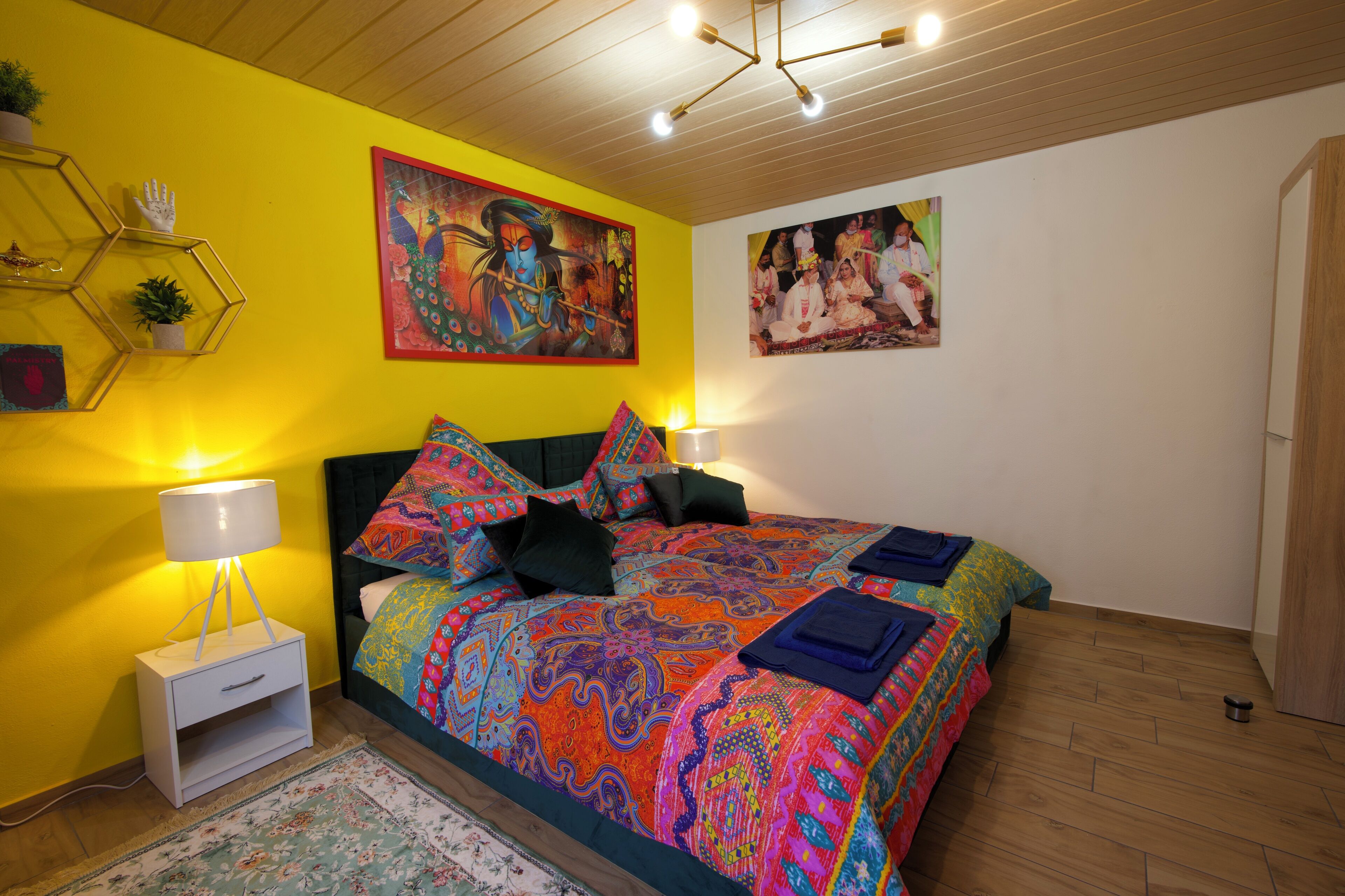A bedroom with a colorful bedspread, a nightstand with a lamp, and two wall-mounted paintings.