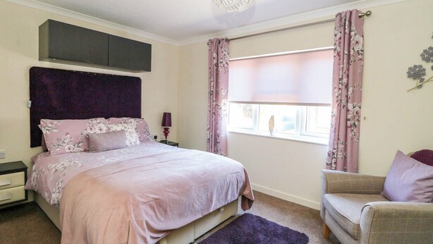 A bedroom with a bed, a nightstand, a dresser, a chair, a window with curtains, and a ceiling light. - Rosegrace Lodge (Auchterarder)
