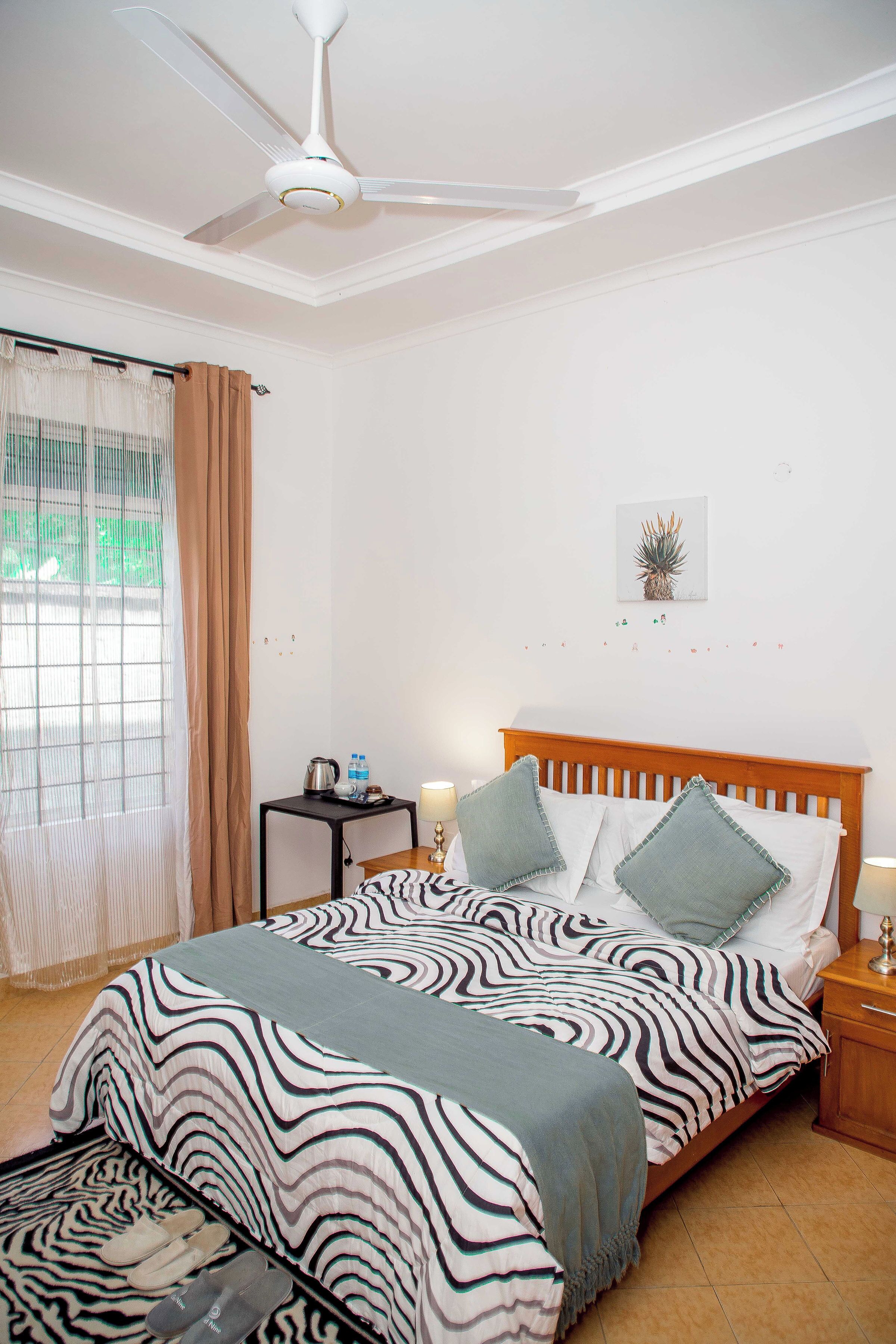 Ein Schlafzimmer mit einem zebra-gestreiften Bettüberzug, einem Deckenventilator, einem kleinen Nachttisch mit Lampe und einem Fenster mit Vorhängen.